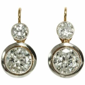 May include: A pair of antique-style drop earrings. Each earring features a large, round, clear gemstone set in a silver-colored bezel with a gold-colored hook. The earrings have a classic, elegant design.