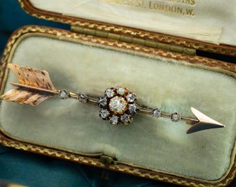 May include: A vintage gold arrow brooch with a central flower design and diamond accents. The brooch is displayed in a velvet-lined box. The arrow's fletching and arrowhead are clearly defined, showcasing the craftsmanship of the jewelry.