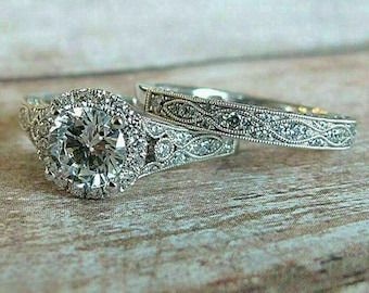 May include: A diamond engagement ring set featuring a large, round-cut center stone surrounded by smaller diamonds. The matching band has an intricate, engraved design. The rings are displayed on a light-colored wooden surface.