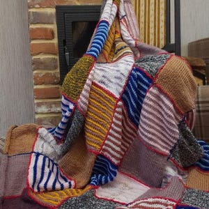 May include: A handmade patchwork blanket with a variety of knitted squares in shades of brown, blue, yellow, and gray. The blanket has a red border and is draped over a chair. The blanket is a cozy home decor item.