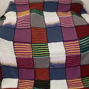 May include: A colorful, patchwork knitted blanket. The blanket features squares of various colors, including white, blue, purple, red, gray, yellow, and green. The blanket is draped over a surface, showcasing its textured design.