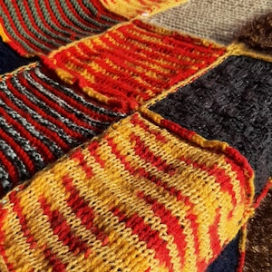 May include: Close-up of a patchwork textile with a variety of knitted squares. The squares feature vibrant colors including red, yellow, gray, and green. The knitted texture is visible, creating a cozy and handcrafted appearance.