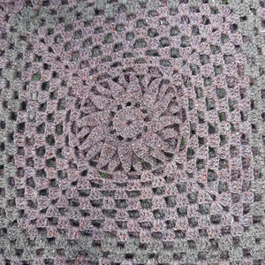May include: Close-up of a handmade crochet item. The piece features a central floral design in shades of purple and grey, surrounded by a square, open-weave pattern. The yarn has a textured appearance, adding depth to the design.