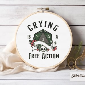 Puede incluir: Un aro de bordado blanco con el texto "Crying is a Free Action" y un dado verde de 20 caras con una calavera y flores.