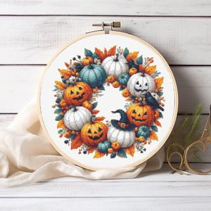 May include: A round embroidery hoop featuring a Halloween-themed cross-stitch design. The design includes orange jack-o'-lanterns, white and teal pumpkins, fall leaves, and a black crow. The hoop is set against a white wooden background.