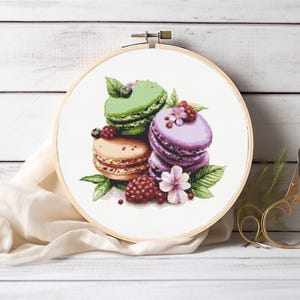 May include: Cross-stitch embroidery of colorful macarons in a wooden hoop. The macarons are green, purple, and tan, decorated with berries and flowers. The embroidery is set against a white wooden background.