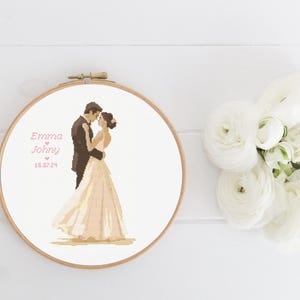 Op de afbeelding: Een witte borduurring met een kruissteekontwerp van een bruid en bruidegom. De tekst "Emma & Johnny 15.07.24" is in roze onder het paar geborduurd.