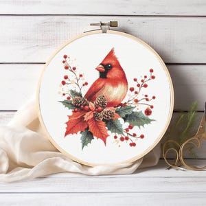 May include: A round embroidery hoop featuring a vibrant red cardinal bird perched amidst holly leaves, berries, and pinecones. The artwork is set against a white background, creating a festive, seasonal theme. A pair of gold scissors rests nearby.
