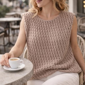 Peut inclure: Un haut sans manches en crochet marron clair et texturé. Le haut a une encolure ronde et une coupe légèrement ample. La personne est assise à une table avec une petite tasse et une soucoupe blanches. L'arrière-plan est un cadre de café extérieur flou.