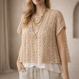 May include: A beige crochet top with short sleeves and a round neckline. The top is layered over a white blouse and paired with beige linen pants. A layered necklace with a pendant adds a touch of elegance to the outfit. The top is made of a soft, open-weave material.