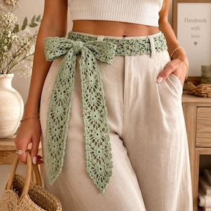 Könnte beinhalten: Ein salbeigrüner Häkelgürtel mit einem komplizierten Blumenmuster und einer langen Schleife. Der Gürtel wird über einer hellbeigen Leinenhose mit hoher Taille getragen. Das Häkeldesign besteht aus sich wiederholenden kreisförmigen Motiven.