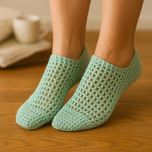 Könnte beinhalten: Ein Paar hellgrüne, offen gewebte Häkelsocken. Die Socken werden an Füßen getragen, mit einem Holzboden im Hintergrund. Die Socken haben ein strukturiertes, gitterartiges Muster und wirken weich und bequem.