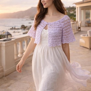 May include: A light purple, open-front crochet shrug with short sleeves. The shrug is worn over a white dress. The background shows a coastal scene with a white balcony and ocean view. The shrug is a delicate, lacy design.