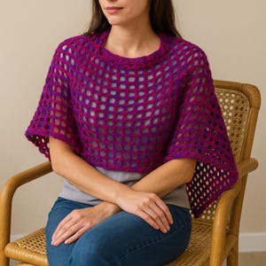 May include: A striking, open-weave poncho in shades of purple and magenta. The crocheted garment has a wide neckline and short sleeves. The poncho is worn over a grey top and blue jeans, creating a casual, layered look.