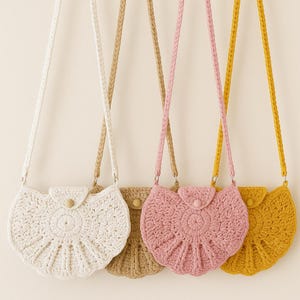 Puede incluir: Cuatro bolsos de crochet en blanco, beige, rosa y amarillo, cada uno con un diseño circular y una correa trenzada. Los bolsos tienen un cierre de solapa con un botón de madera. Los bolsos cuelgan sobre un fondo neutro.