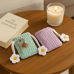 May include: Two handmade crochet key pouches, one mint green and one lavender, each with a white drawstring and a daisy accent. The mint pouch holds keys and coins. A lit candle and a book are in the background.