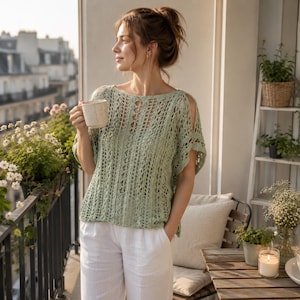 May include: A light green, open-weave crochet top with short sleeves and a scoop neckline. The top is paired with white linen pants. The image is taken on a balcony with plants and a cup of coffee.