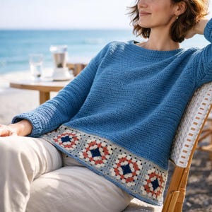 May include: A blue crochet sweater with a decorative border featuring red, white, and navy blue squares. The sweater has long sleeves and a round neckline. The person is sitting in a chair near the ocean.