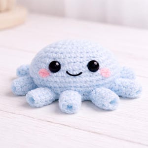 May include: A light blue crocheted octopus with eight tentacles. The octopus has large black eyes, pink cheeks, and a smiling mouth. The handmade plush toy is sitting on a white wooden surface.