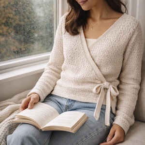 Può includere: Maglione avvolgente color crema con maniche lunghe e chiusura con fiocco. Il maglione ha un motivo a maglia strutturato e uno scollo a V. La persona indossa jeans azzurri e tiene in mano un libro aperto.