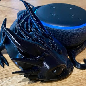 Peut inclure: Une figurine de dragon noire imprimée en 3D tenant un haut-parleur intelligent Amazon Echo Dot noir.