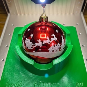 Op de afbeelding: Rode kerstbal met een gouden dop, met een wit ontwerp van de kerstman in een slee met rendieren, een huis en een kerstboom. De bal heeft een diameter van 60 mm en ligt in een groene houder.