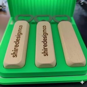 Op de afbeelding: Drie flesopeners met houten handvatten en groene metalen koppen worden gepresenteerd in een groene plastic koffer. De tekst "shiredesignco" staat op twee van de houten handvatten. De koffer is open en onthult de openers.