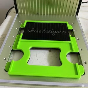 May include: A laser engraving machine with a bright green frame and a black insert. The text "shiredesignco" is visible on the black insert. The machine is open, revealing the interior components.