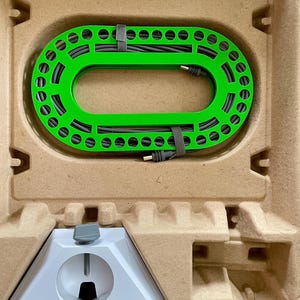 May include: A bright green, oval-shaped device with evenly spaced holes and a black cable, secured with a grey strap, sits within a cardboard container. Below, a white, angular support structure with a grey knob is visible.