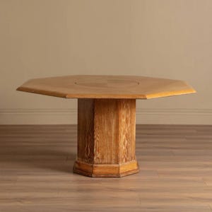 Può includere: Tavolo ottagonale in legno marrone chiaro, con base a piedistallo. Il piano del tavolo ha un incavo circolare. Il tavolo è in legno chiaro e poggia su un pavimento in legno. Le sue dimensioni sono di circa 120 cm di diametro.