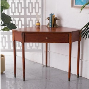 May include: A mid-century modern, hexagonal-shaped wooden table with a single drawer and brass-tipped legs. The table is a rich, warm brown color and features a small, round drawer pull. The table is approximately 30 inches tall.