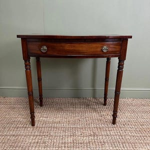 Op de afbeelding: Een mahoniehouten, halfronde bijzettafel met een enkele lade en messing hardware. De tafel heeft vier slanke, gedraaide poten. De tafel is ongeveer 76 cm breed en 76 cm hoog.