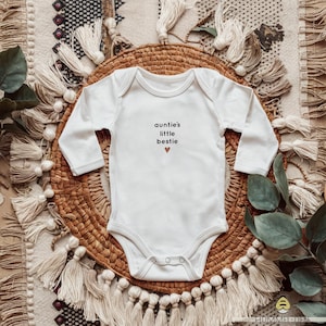 以下が含まれることがあります： 「auntie's little bestie」という文字と小さなハートがプリントされた白い長袖のベビーロンパースです。