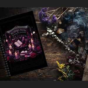 Peut inclure: Un cahier &agrave; spirales noir avec l'inscription "Reading Filth & Minding My Business" et "The Book Club" sur la couverture. L'illustration de la couverture comprend des bougies, des roses et un livre ouvert. L'image est pos&eacute;e sur une surface en bois avec des &eacute;l&eacute;ments d&eacute;coratifs.