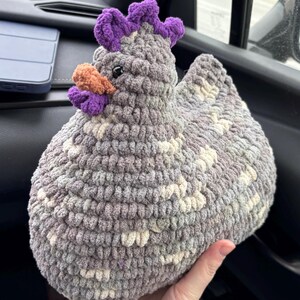 May include: A crocheted gray and white chicken plush toy with a purple comb and beak. The toy is being held in a person's hand.