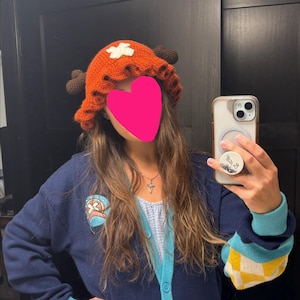 May include: A crocheted orange bucket hat featuring a ruffled brim, dark brown antlers on the sides, and a white cross emblem on the front. The hat is paired with a navy blue cardigan that has a light blue and yellow checkered pattern on the sleeve.