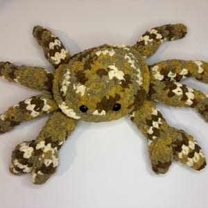 May include: A large, handmade crochet crab plush toy. The crab has a round body and eight legs in shades of brown, green, and cream. The crab has two black button eyes. The plush is designed to be soft and cuddly.