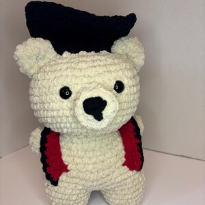 Pode incluir: Um urso de crochê feito à mão, de cor creme, usando um chapéu preto e suspensórios vermelhos e pretos. O urso tem olhos de botão pretos e um nariz preto. O urso está em pé e é feito de um fio macio e texturizado.
