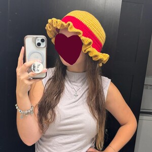 Op de afbeelding: Een handgehaakte bucket hat met een felgele kroon, een brede, geplooide gele rand en een opvallende horizontale rode band rond de basis van de kroon. De hoed is volledig gemaakt met een gestructureerd garenpatroon in een haaksteek.