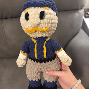 May include: Handmade crochet doll with a blue cap, navy blue shirt with yellow accents, and tan pants. The doll has a cream-colored face with a yellow mustache and black eyes. The doll is being held by a person.