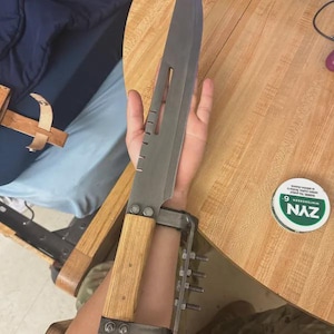 May include: A large, custom-made knife with a wide, gray blade and a light-colored wooden handle. The knife is attached to a metal arm brace with multiple screws. The image is taken from a close-up perspective.