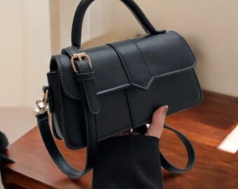 May include: A black handbag with a top handle and a detachable shoulder strap. The rectangular bag features a flap closure with a gold-coloured buckle. The bag is held by a person.