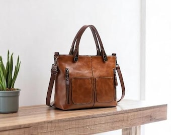 May include: A brown leather tote bag with two front pockets, zippers, and a shoulder strap. The bag has two top handles and is sitting on a wooden surface. The bag is a medium brown colour.
