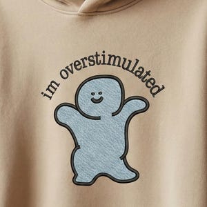 May include: A beige hooded sweatshirt with a drawstring and a front pocket. It displays a blue cartoon character with the text "im overstimulated" in black. The hoodie is made of a soft material, suitable for casual wear.