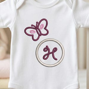 Puede incluir: Body de bebé blanco con una mariposa y la letra "H" bordadas en tonos morados. La mariposa está encima de un círculo que contiene la letra "H". El body se muestra en una cuna blanca.