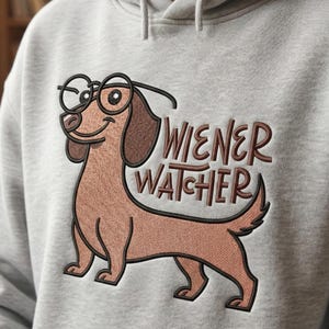 Op de afbeelding: Een lichtgrijze hoodie met een geborduurd cartoonteckel met een bril. De teckel is bruin met een lichtbruine buik. De tekst "WIENER WATCHER" is bruin geborduurd. Op de achtergrond is een echte teckel te zien.