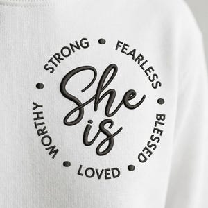 May include: White sweatshirt with the embroidered phrase "She is" in a circular design. The words "Strong", "Fearless", "Blessed", "Loved", and "Worthy" are embroidered around the circle in black.