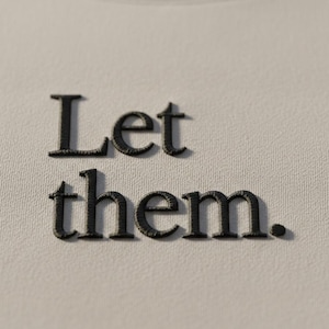 May include: Close-up of a light gray sweatshirt with the embroidered words "Let them." in black thread. The text is centered and the fabric has a subtle texture.