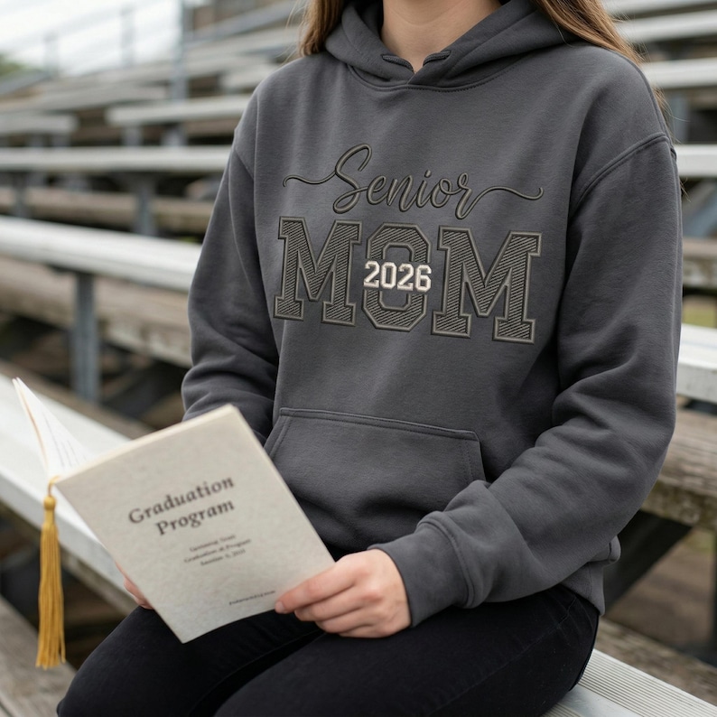 Lifestyle embroidery mockup of a "Senior Mom 2026" design on a gray hoodie. A model is shown sitting on stadium bleachers holding a blurred graduation program, emphasizing the realistic thread shine, stitch density, and authentic raised texture of the embroidery.
