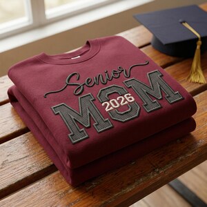 A maroon sweatshirt with a "Senior Mom 2026" embroidered logo, neatly folded on a wooden bench. A graduation cap is visible in the blurred background, showcasing the detailed fill stitching and natural window lighting on the premium fabric.